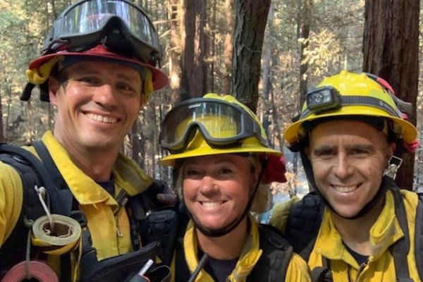 Three firefighters smiling at the camera and wearing yellow wildland gear with trees behind them.